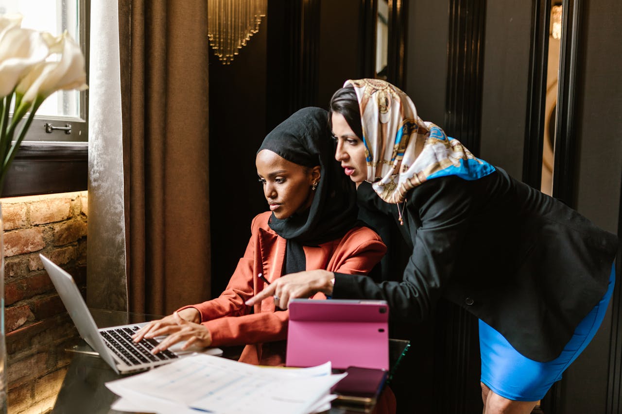 Two women in business attire collaborate at a laptop indoors, focusing on teamwork and technology.