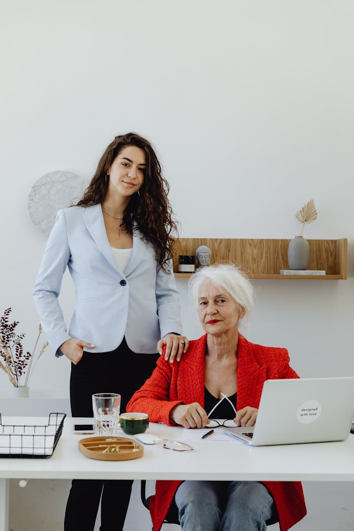 Two businesswomen, one senior, collaborating at a desk in a modern office with laptops and documents.