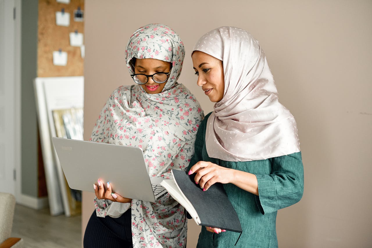 Two Muslim women in hijabs collaborate on a project using a laptop and notebook in an office setting.